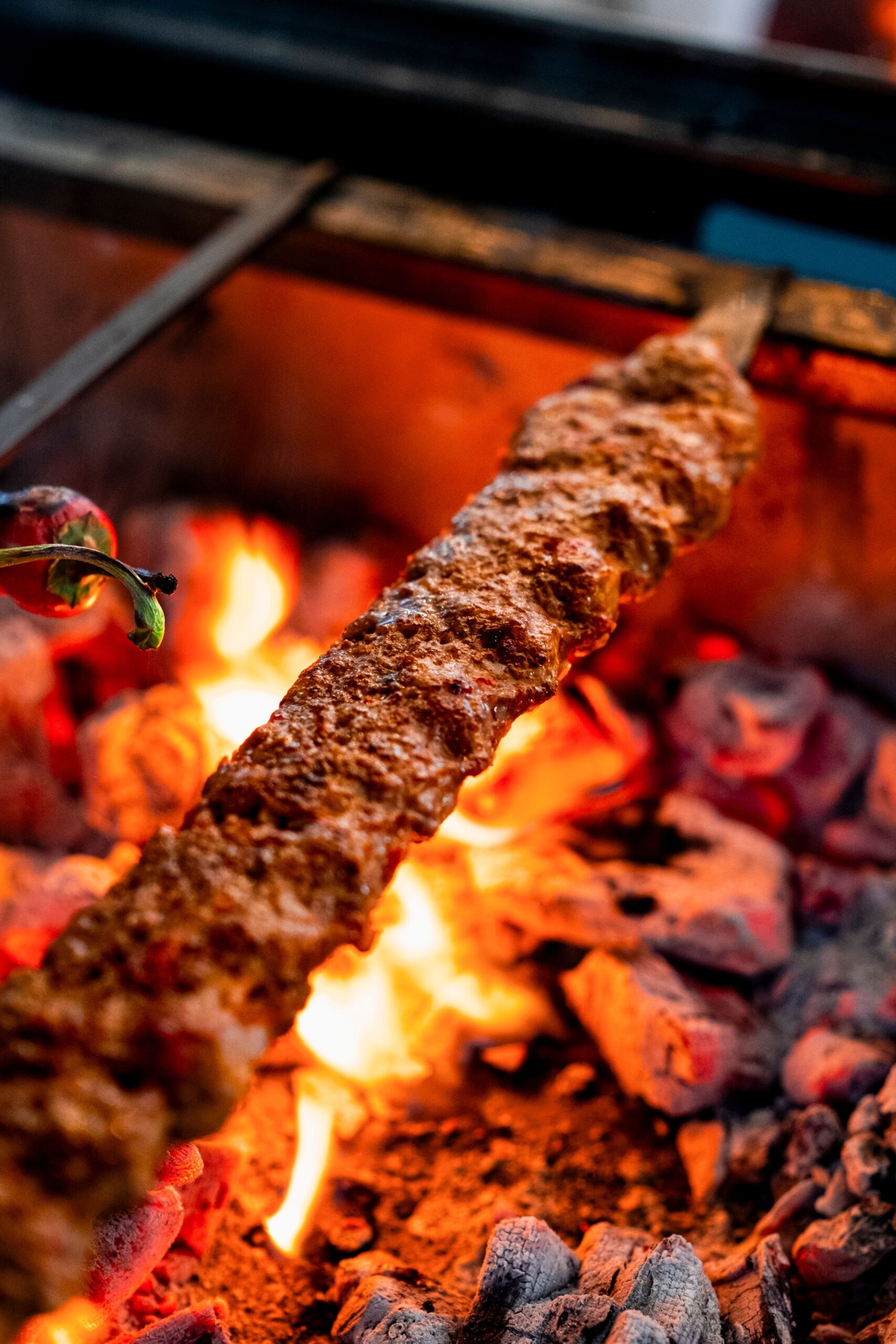 Close-up of spicy Adana kebab grilling over hot charcoal with flames.